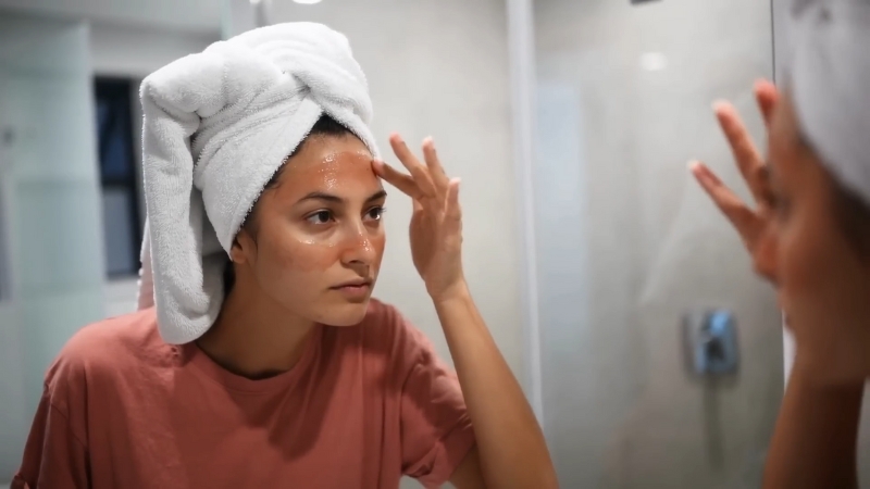 Woman with a towel on her head applying skincare while looking at her reflection in a bathroom mirror