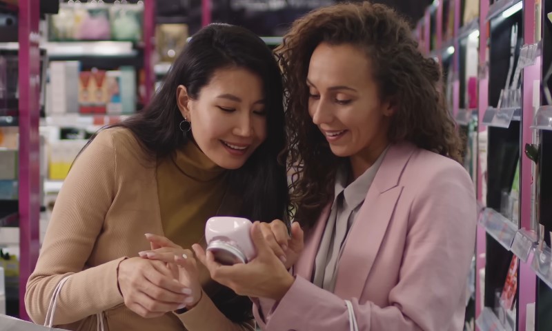Two women shopping in a beauty store, smiling and examining a pink product