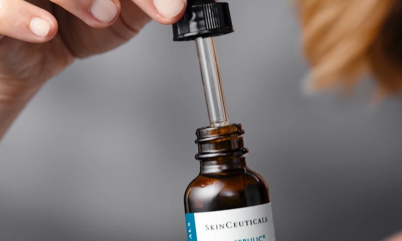 A hand holds a dropper above an open brown skincare bottle labeled SkinCeuticals